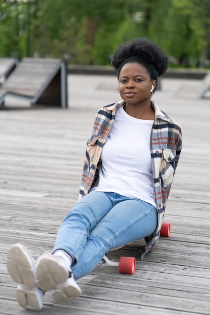 Une fille africaine décontractée et décontractée se détend après avoir fait de la planche à roulettes dans un skatepark, assise sur un longboard, écoutez de la musique à l'aide d'un casque sans fil. Femme noire calme jeune adulte en jeans à l'extérieur dans un parc urbain