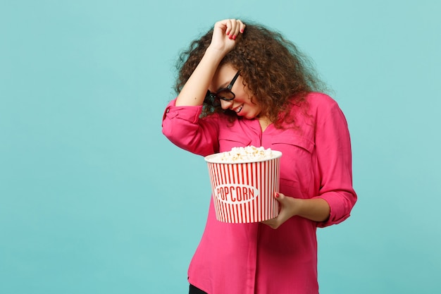 Fille africaine effrayée dans des lunettes imax 3d regardant un film de film tenir un revêtement de pop-corn avec une main isolée sur fond de mur bleu turquoise. Émotions des gens au cinéma, concept de style de vie. Maquette de l'espace de copie.