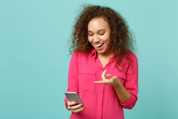 Fille africaine excitée dans des vêtements décontractés roses à l'aide d'un téléphone portable, tapant un message sms isolé sur fond de mur bleu turquoise en studio. Les gens émotions sincères, concept de style de vie. Maquette de l'espace de copie.