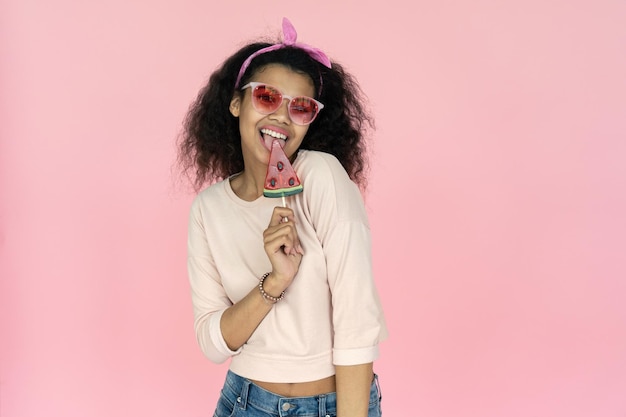 Une fille africaine heureuse en lunettes lèche des bonbons de pastèque isolés sur un fond rose.