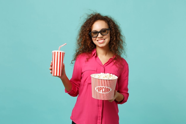 Une fille africaine souriante dans des lunettes imax 3d regardant un film de film tenir du pop-corn, une tasse de soda isolée sur fond bleu turquoise en studio. Émotions des gens au cinéma, concept de style de vie. Maquette de l'espace de copie.