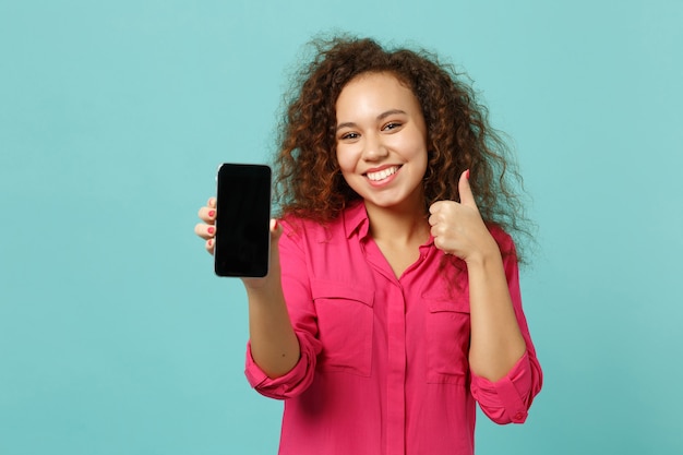 Une fille africaine souriante dans des vêtements décontractés montrant le pouce vers le haut tient un téléphone portable avec un écran vide vierge isolé sur fond bleu turquoise. Les gens émotions sincères, concept de style de vie. Maquette de l'espace de copie.