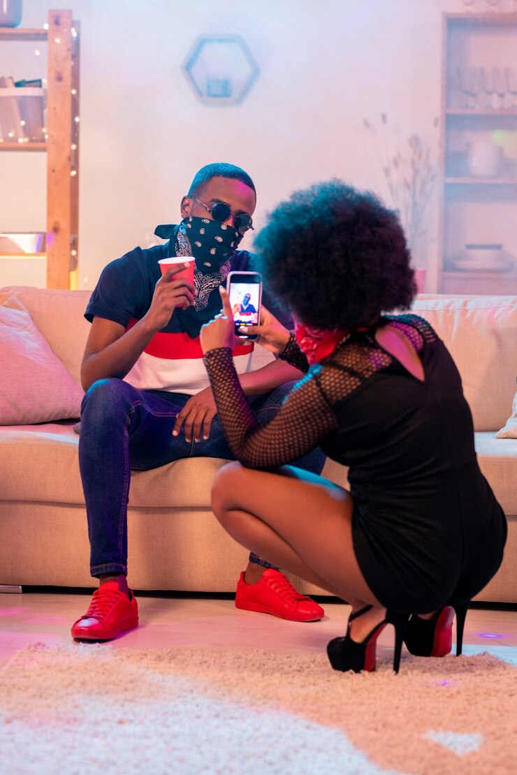 Fille africaine en tenue noire assise sur des squats et prendre une photo de son petit ami avec un verre assis sur un canapé moelleux dans le salon