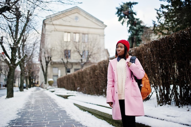 Fille Afro-américaine Au Chapeau Rouge Et Manteau Rose Avec Sac à Dos Dans La Rue De La Ville Le Jour De L'hiver