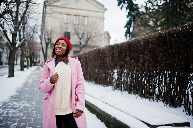 Fille afro-américaine en chapeau rouge et manteau rose dans la rue de la ville le jour de l'hiver.