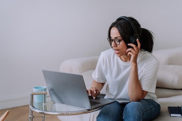 Une fille afro-américaine concentrée en t-shirt blanc et des lunettes assises sur un canapé travaille à l'aide d'un ordinateur portable à la maison