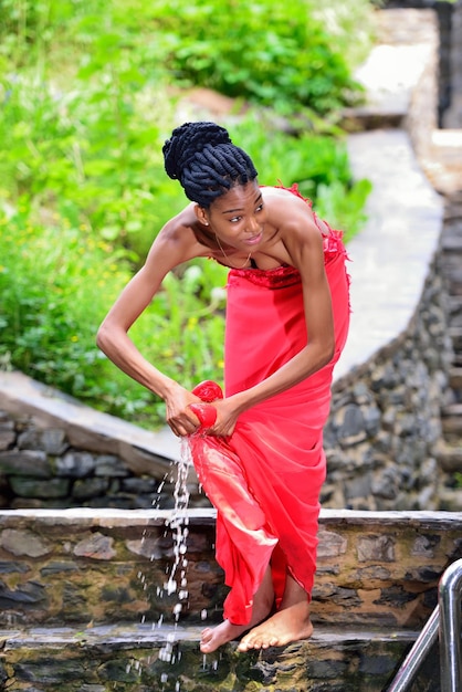 Une fille afro-américaine avec des dreadlocks se tient dans la nature au bord de l'eau et lave une robe rouge