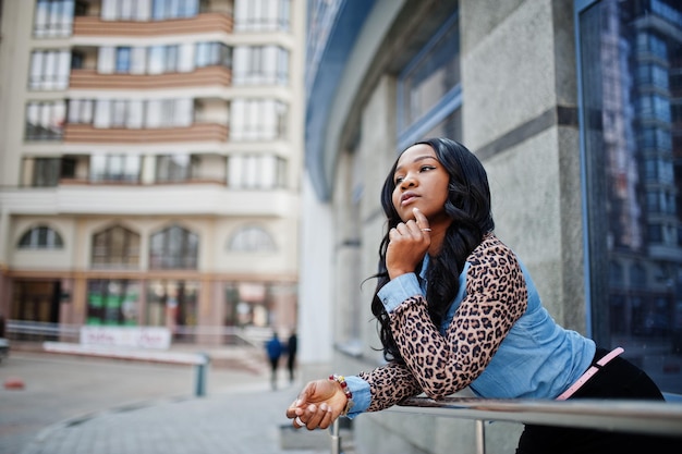 Fille afro-américaine hipster portant une chemise en jean à manches léopard posant dans la rue contre un immeuble de bureaux moderne avec des fenêtres bleues