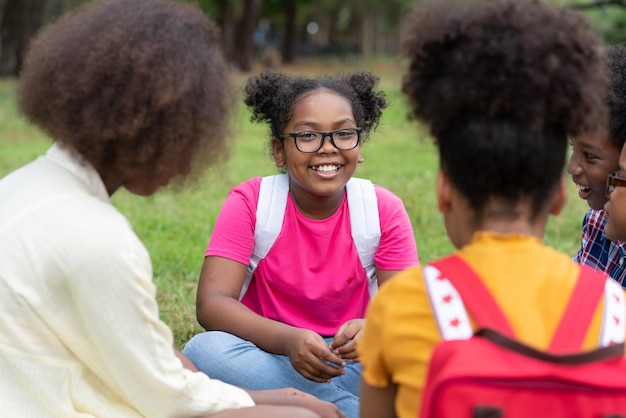 Fille afro-américaine avec des sacs à dos et des enfants assis sur l'herbe dans le parc