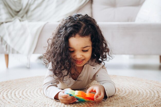 Photo fille afro-américaine sur le sol à la maison jouant avec un jouet pop en appuyant sur des boutons soulageant le stress
