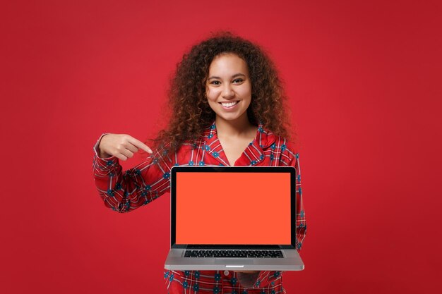 Photo une fille afro-américaine souriante en pyjama homewear reste à la maison isolée sur fond rouge. détendez-vous le concept de mode de vie de bonne humeur. maquette de l'espace de copie. pointant sur un ordinateur portable avec un écran vide vide.