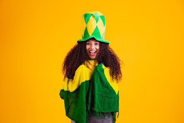 Fille afro encourageant l'équipe brésilienne préférée, tenant le drapeau national sur fond jaune.