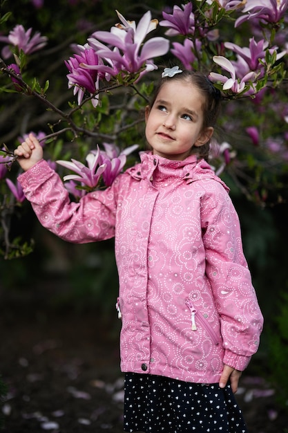 Fille d'âge préscolaire en veste rose profitant d'une belle journée de printemps près de l'arbre en fleurs de magnolia Activités de printemps