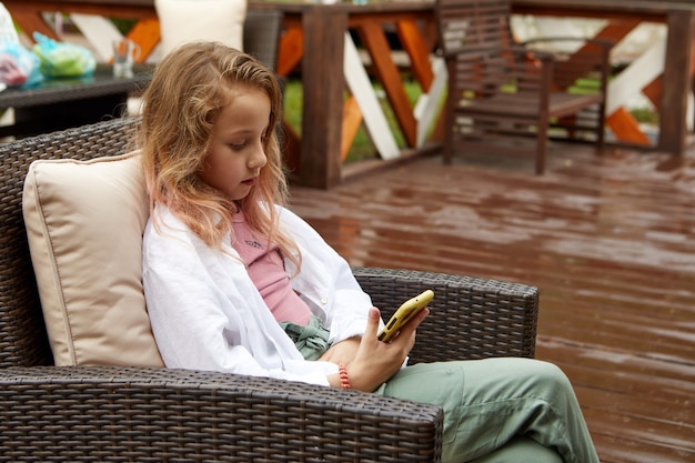 Fille à l'aide de smartphone n fauteuil en osier sur terrasse