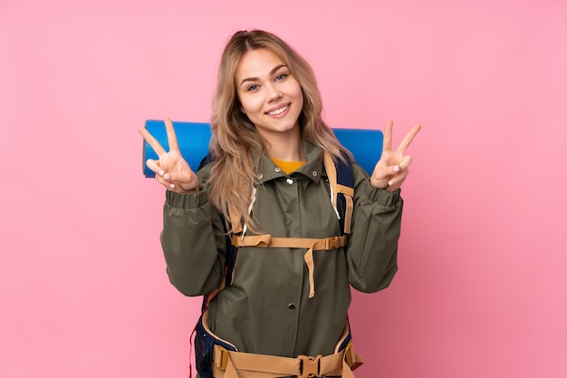 Fille d'alpiniste adolescent avec un gros sac à dos sur le mur rose montrant le signe de la victoire avec les deux mains