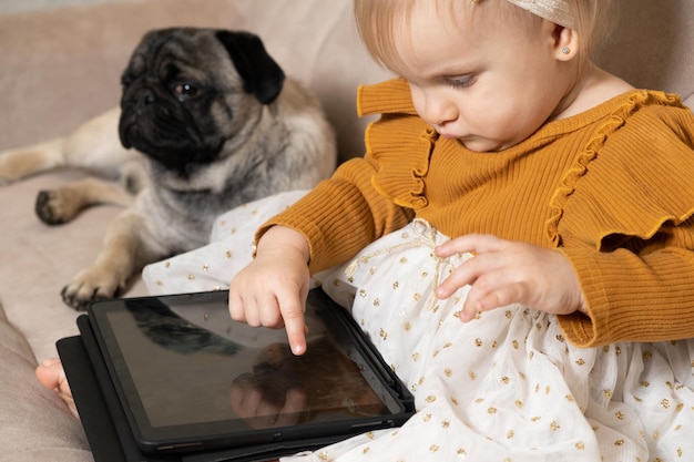 Une fille d'un an avec un carlin regarde des dessins animés sur une tablette assise sur le canapé Gadgets pour les enfants à l'écran