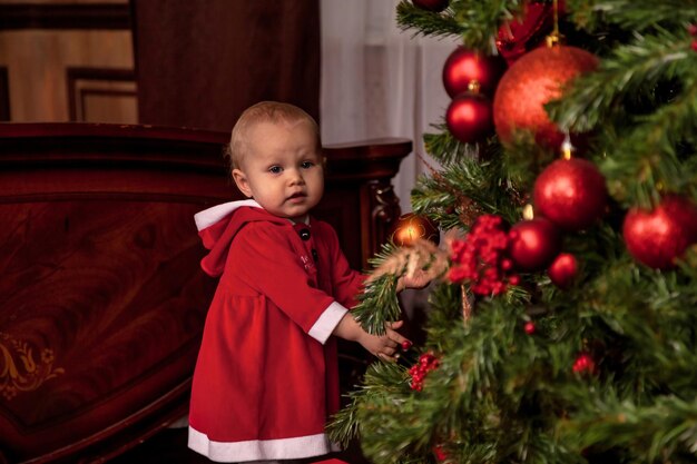 Une fille d'un an déguisée en Père Noël sur fond d'arbre de Noël décoré à l'intérieur en regardant la caméra