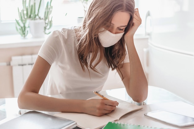 Une fille apathique et dépressive dans un masque médical travaille à distance à la maison et s'assoit à table, écrit dans un cahier. Fille étudiante étudiant à la maison, isolement de quarantaine, concept de séjour à la maison