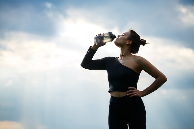 Une fille d'apparence européenne avec une carrure sportive dans un haut noir et des leggings noirs sur un ciel bleu. Elle boit de l'eau à la bouteille. Mode de vie sain, fille brune athlétique. Motivation pour le sport