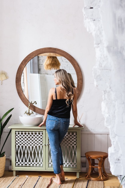 Une fille d'apparence européenne se tient près d'une table avec un miroir et prenadlezhnostyami pour le maquillage, le parfum, l'évier, vêtu d'un jean