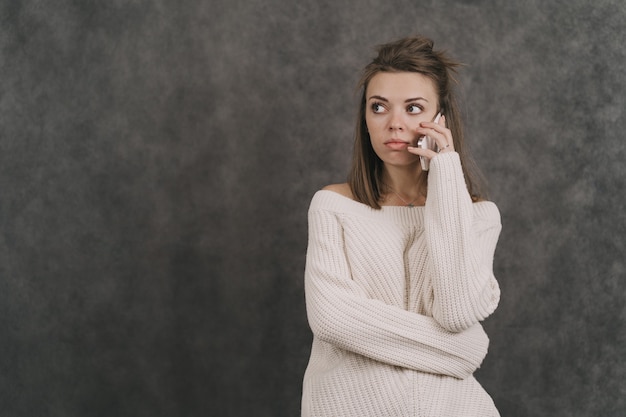 La fille appelle au téléphone. Une fille dans un pull blanc sur un pull gris. La fille a une conversation sérieuse au téléphone.