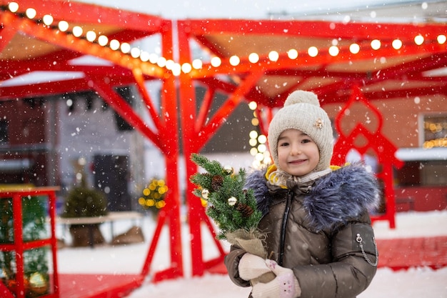 Une fille avec un arbre de Noël dans ses mains en plein air dans des vêtements chauds en hiver sur un marché festif Des guirlandes de lumières féeriques décorées de la ville de neige pour la nouvelle année