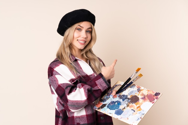 Photo fille artiste adolescent tenant une palette isolée sur bleu pointant vers l'arrière