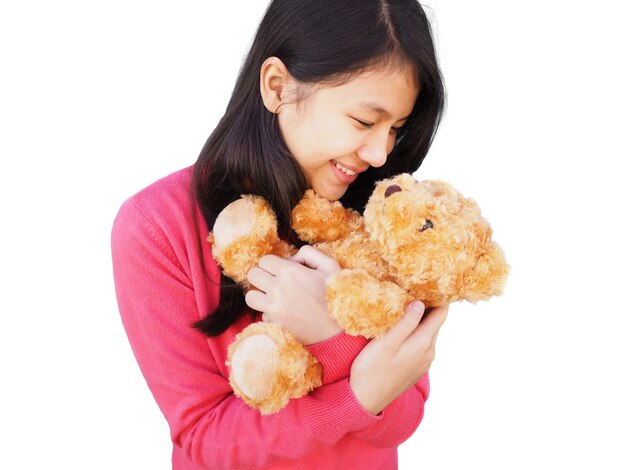 Fille asiatique aux cheveux longs portant une chemise rose et souriant à l'ours en peluche brun dans son bras hug holding, isolé