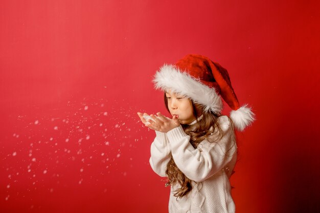 Fille asiatique en bonnet de noel soufflant sur la neige sur fond rouge