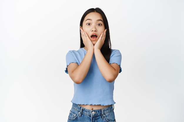 Une fille asiatique choquée haletant en regardant la caméra a surpris en vérifiant quelque chose de grande nouvelle debout sur fond blanc