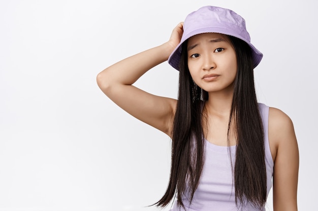 Une fille asiatique confuse se gratte la tête et a l'air perplexe, ne peut pas comprendre quelque chose, debout dans un chapeau de seau et un débardeur sur blanc.