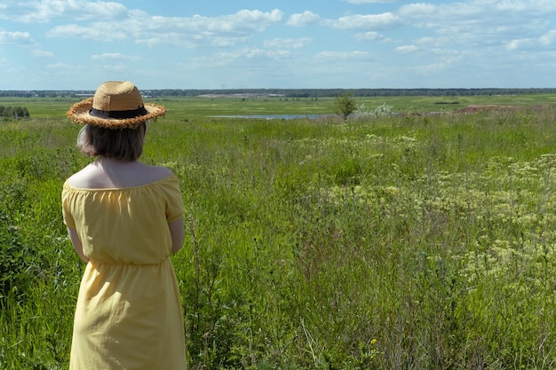 Fille asiatique dans un chapeau de paille et une robe jaune parmi l'herbe duveteuse dans le champ vue arrière Heure d'été