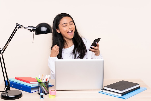 Fille Asiatique Dans Un Lieu De Travail Avec Un Ordinateur Portable Sur Un Mur Beige Avec Téléphone En Position De Victoire