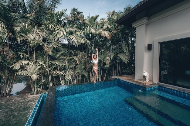 Fille asiatique debout au bord de la piscine, ses bras au-dessus de sa tête. Territoire d'un hôtel exotique, palmiers et eau bleue ; relaxation.