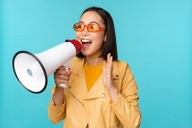 Fille asiatique élégante faisant une annonce dans un mégaphone criant avec haut-parleur et souriant invitant les gens à recruter debout sur fond bleu