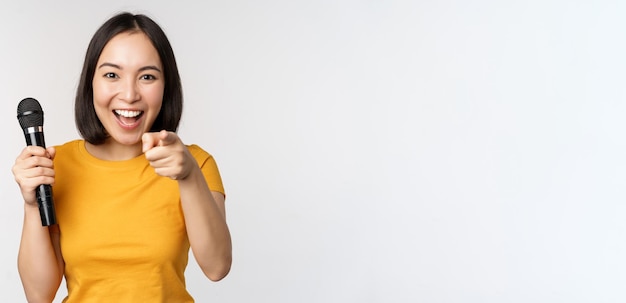 Fille asiatique enthousiaste avec microphone pointant du doigt la caméra vous suggérant de chanter debout sur fond blanc