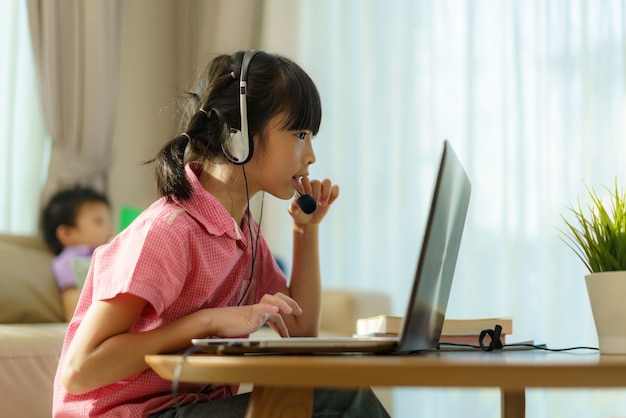 Fille asiatique étudiant par vidéoconférence e-learning avec l'enseignant et ses camarades de classe sur ordinateur et son livre de lecture de frère dans un canapé dans le salon à la maison