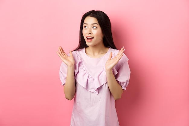 Fille asiatique excitée applaudit et regarde à gauche, regardant la performance et les applaudissements, debout en robe sur fond rose.