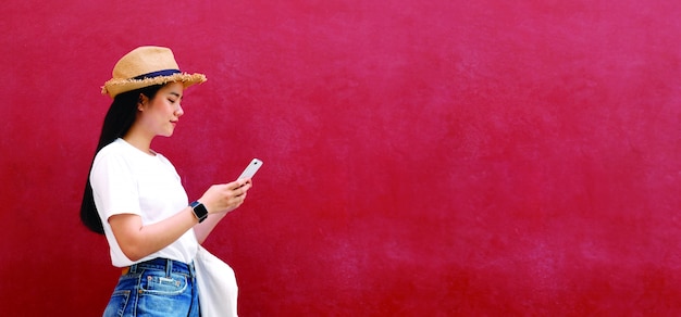 Fille asiatique heureuse à l'aide de téléphone portable debout sur fond de mur rouge