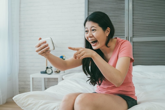 une fille asiatique heureuse assise au chevet en vêtements décontractés pointe vers l'écran du téléphone tout en regardant une vidéo en ligne amusante dans la chambre à la maison pendant la journée.