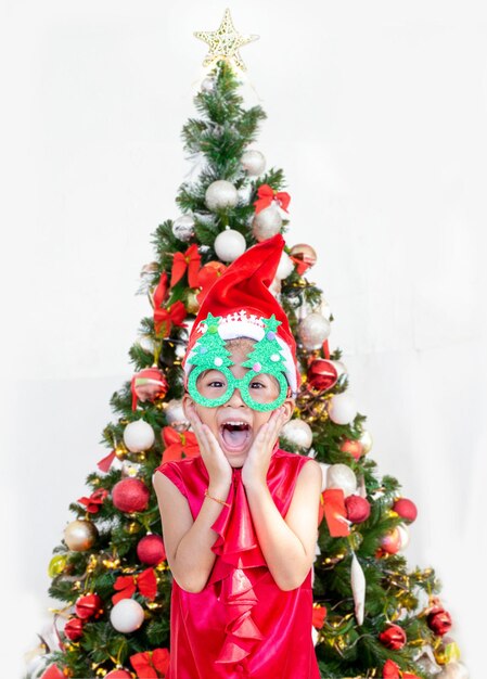 Photo fille asiatique heureuse enfant avec robe rouge de père noël action surprise avec décoration d'arbre de noël xmas et nouvelle année concept saisonnier pour la publicité