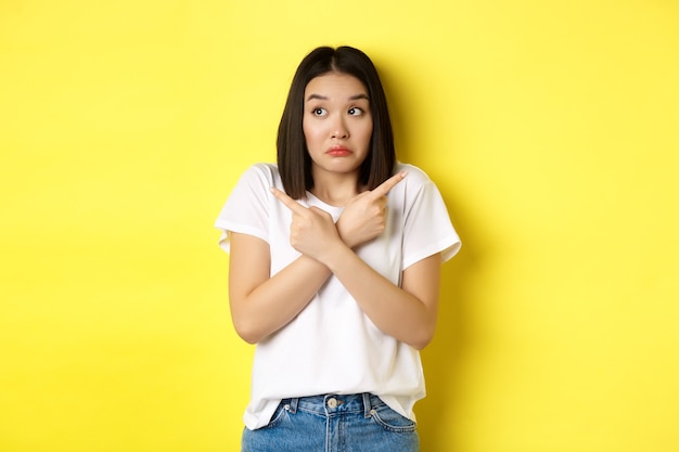 Une fille asiatique indécise a besoin d'aide pour choisir, pointant les doigts sur le côté et ayant l'air confus, debout sur fond jaune.