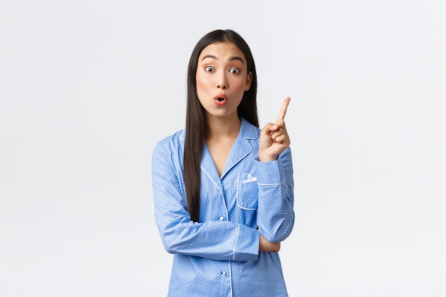 Fille asiatique intelligente et réfléchie en pyjama bleu levant l'index pour suggérer un plan, avoir une idée, être frappé d'une grande réflexion. Femme en solution composée de jammies, mur blanc debout