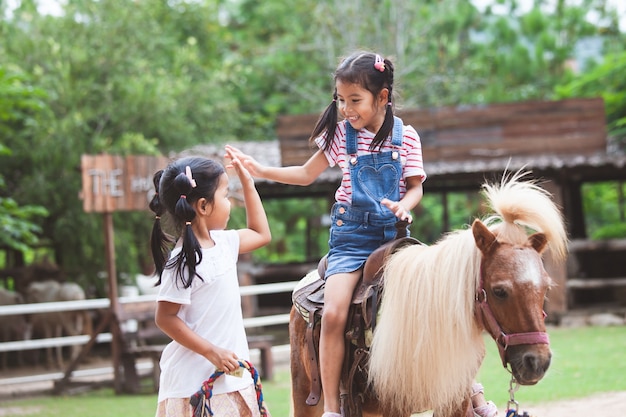 Fille asiatique mignon enfant chevauchant un poney et faisant salut cinq geste avec sa soeur aînée