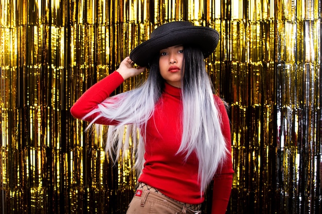 Une fille asiatique à la peau bronzée porte un chapeau foncé avec des cheveux blancs dans l'air, tourne les sourires et se sent heureuse danser sur une fête de rideau en feuille d'or avec plaisir, éclairage de studio sur fond doré, espace de copie