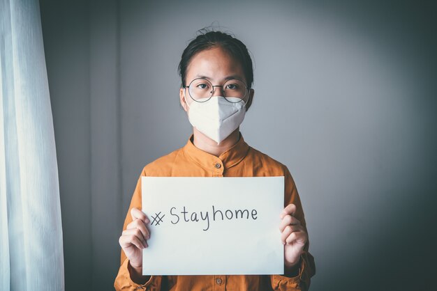 Une fille asiatique portant des lunettes, portant un masque, tenant du papier étiqueté #stayhome, s'ennuie de devoir détenir et traiter la maladie seule à la maison. Concept de quarantaine à domicile, prévention COVID-19