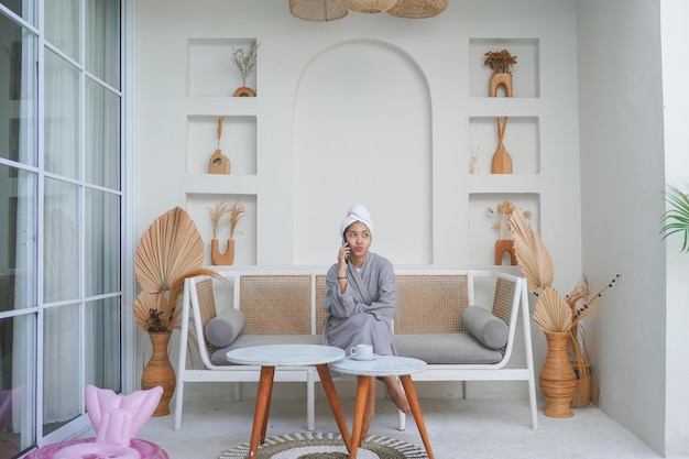 Une fille asiatique réfléchie en peignoir gris assise sur un canapé en train de penser et de converser au téléphone.