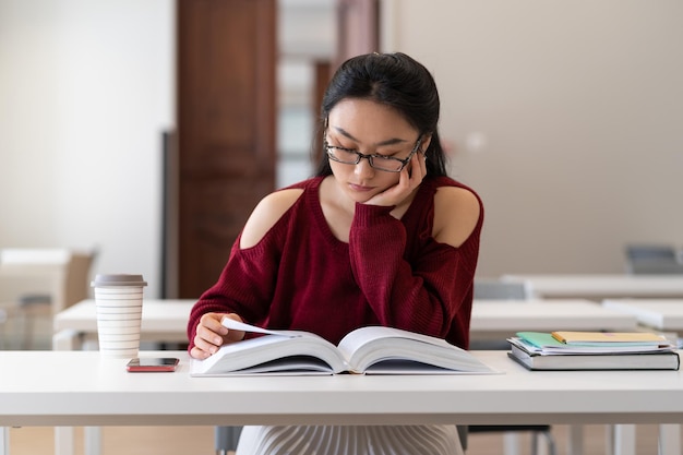 Fille asiatique ringard lisant un manuel tout en se préparant à un examen ou à un test dans la salle de lecture de la bibliothèque