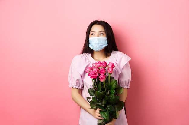 Fille asiatique romantique dans un masque médical regardant de côté un espace vide avec des yeux rêveurs, tenant un bouquet de roses pour la Saint Valentin, ayant rendez-vous avec son amant pendant la pandémie.
