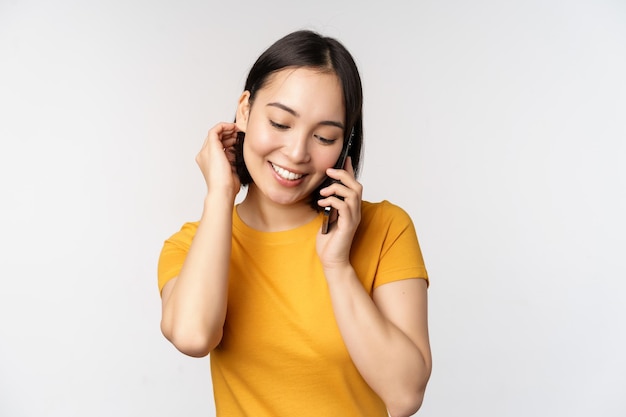 Une fille asiatique romantique et mignonne répond à un appel téléphonique en parlant sur un mobile souriant coquette debout en t-shirt jaune sur fond blanc